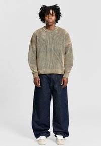 Maglione khaki a righe con polsini a costine, abbinato a jeans blu scuro a vestibilità larga e sneakers bianche. Presenta una silhouette rilassata e texture.
