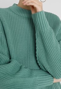 Strukturierter, gestrickter grüner Pullover mit hohem Hals, weiten Bündchen und fallenden Schultern. Verfügt über ein dezentes gewebtes Muster im gesamten Design.