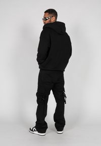 Haut à capuche noir avec une grande capuche, associé à un pantalon cargo noir avec des poches latérales et des accents en métal. Les chaussures comprennent des baskets noires et blanches.