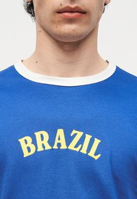 Κοντινό πλάνο ενός ατόμου που φορά μπλε πουκάμισο με λευκό γιακά, με τη λέξη «BRAZIL» σε μεγάλα κίτρινα γράμματα στο στήθος.