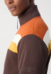 Hochkragen-Sweatshirt in Braun, Orange, Weiß und Gelb mit Farbkombination aus strukturiertem Stoff; verfügt über ein Logo am Bündchen und einen Reißverschluss.