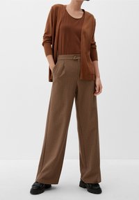 Hnědý cardigan přes hnědý top, kombinovaný s širokými kalhotami s kostkovaným vzorem v odstínech hnědé a béžové. Černé boty s třásňovými prvky.