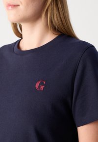 Femme portant un t-shirt bleu marine avec une petite lettre "G" rouge brodée sur la poitrine gauche.