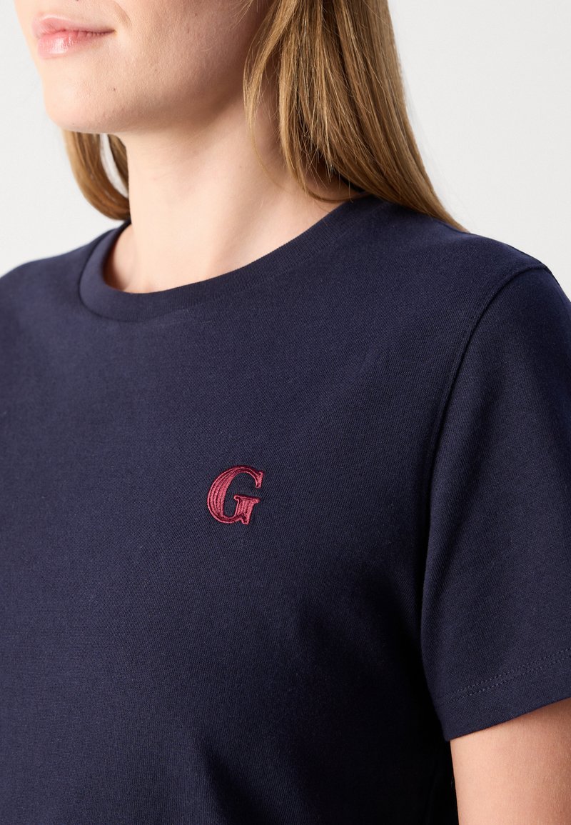 Frau trägt ein navyblaues T-Shirt mit einem kleinen roten gestickten Buchstaben "G" auf der linken Brust.