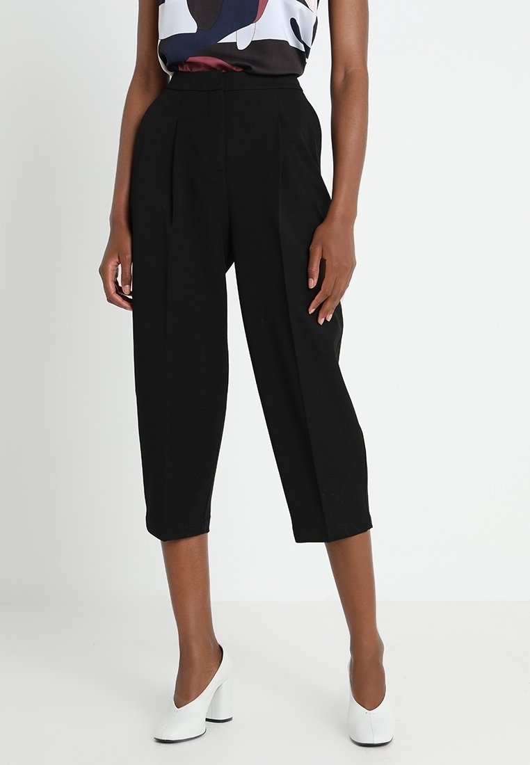 Svarta beskurna byxor med veck, hög midja, skräddarsydd passform, slät textur. Modellen bär vita klackar, tillsammans med en mönstrad topp.