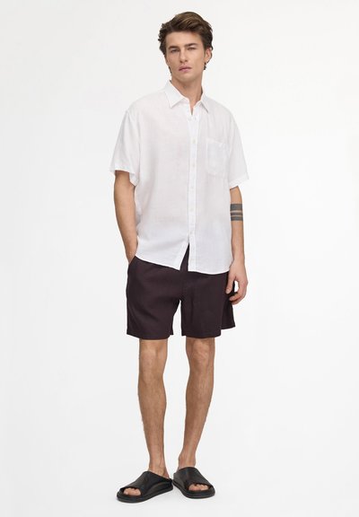 Jeune homme debout, les mains dans les poches, portant une chemise blanche à manches courtes boutonnée, un short noir, des sandales noires et un tatouage sur le bras.