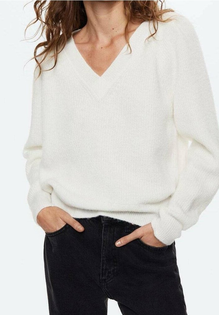 Femme portant un pull blanc doux à col en V avec manches longues, les mains dans les poches d'un jean noir taille haute sur un fond uni.