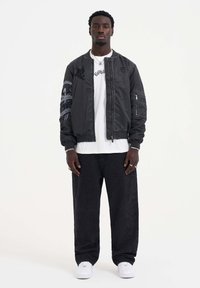 Black bomber jacket with graphic details on the sleeve, paired with a white t-shirt and black wide-leg jeans. White sneakers complete the look.