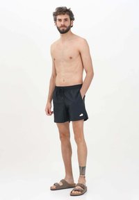 Pantaloni da bagno neri con cintura elastica e tasche laterali. Indossati con sandali marroni. Tessuto liscio e design semplice. Senza motivi o dettagli.