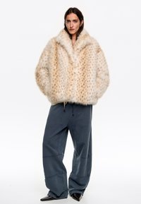 Abrigo de piel sintética crema con un patrón de manchas, silueta oversized y cuello ancho, combinado con pantalones holgados gris pizarra y zapatos negros de punta.