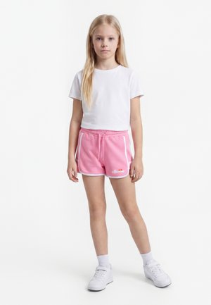 Jeune fille debout, portant un t-shirt blanc, un short rose à cordon avec bordure blanche, des chaussettes blanches et des baskets blanches sur un fond uni.