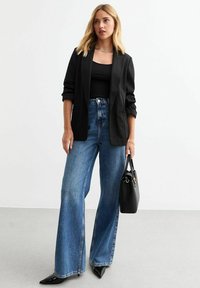 Blazer negro con mangas remangadas, combinado con una camiseta negra sin mangas y unos vaqueros de pierna ancha de cintura alta en azul. Tacones negros de punta y bolso.