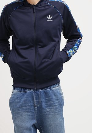 Person trägt eine marineblaue Adidas-Jacke mit Reißverschluss und gemusterten Streifen an den Ärmeln sowie lässige hellblaue Jeans, Hände in den Taschen.