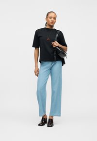 Γυναίκα με πλεξούδες, που φοράει μαύρο T-shirt, ανοιχτό μπλε τζιν με φαρδιά μπατζάκια, μαύρα παπούτσια με τακούνι, κολιέ ροζ και κρατά μαύρη τσάντα ώμου.