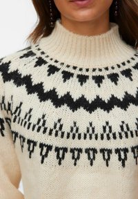 Nahaufnahme eines cremefarbenen Strickpullovers mit schwarzen geometrischen Mustern im Brustbereich und einem hohen gerippten Kragen.