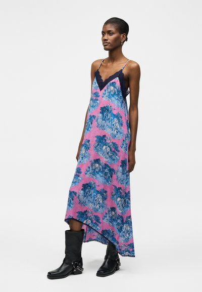 Γυναίκα με ροζ και μπλε μακρύ φόρεμα με λουλουδάτο σχέδιο, διακοσμημένο με σκούρες μπλε δαντέλες, και μαύρα μποτάκια μέχρι τη μέση της γάμπας με αλυσίδα, στέκεται απέναντι σε λευκό φόντο.