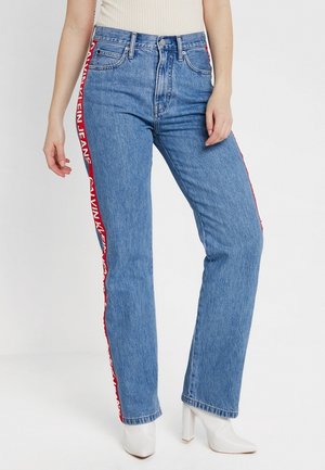 Vrouw die blauwe straight-leg jeans draagt met rode zijstrepen waarop de tekst "Calvin Klein Jeans" staat, gecombineerd met witte enkellaarsjes met hak.