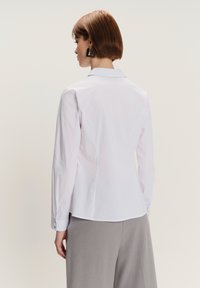 Donna con capelli corti castani che indossa orecchini geometrici neri, camicia bianca con bottoni e pantaloni grigi a gamba larga, voltata di spalle.