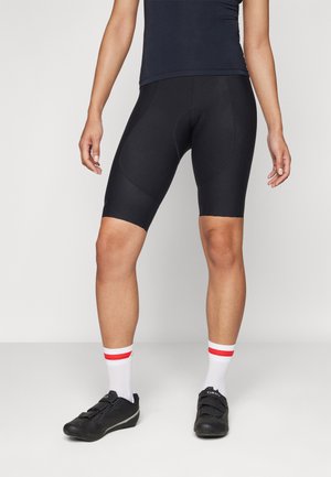 Schwarze Radhosen mit einer enganliegenden Passform, ausgestattet mit gepolsterten Bereichen. Getragen mit schwarzen Radschuhen und weißen Socken mit roten Streifen.