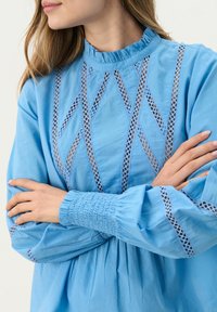 Blusa di cotone blu con scollatura arricciata, motivi a diamante ritagliati e maniche gather con bordo in pizzo decorativo. Finitura tessurizzata.