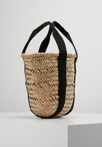 Borsa a secchiello intrecciata in paglia con ampi manici in tessuto nero che si estendono dalla parte superiore alla base, esposta su un piedistallo bianco su uno sfondo neutro.