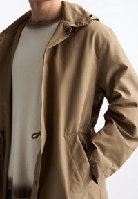 Veste imperméable marron clair avec capuche, boutons sur le devant, taille à cordon de serrage et poches latérales, portée par-dessus un pull gris clair.