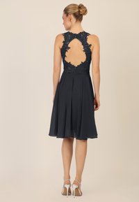 Zwarte mouwloze jurk met kantdetails, open rug en een vloeiende rok. Vergezeld van zilveren strappy hakken.