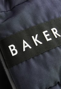 Section en tissu noir avec une texture en grille et une étiquette blanche "BAKER" bien visible, présentant des bords lisses et une fermeture éclair.
