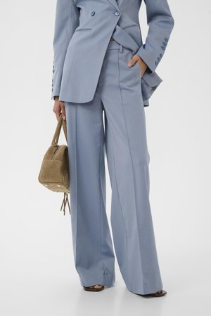 Kobieta w jasnoniebieskich spodniach o szerokich nogawkach i pasującej marynarce, trzyma jasno-brązową zamszową torebkę z frędzlami.