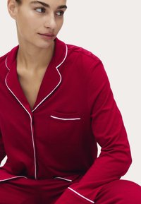 Rød bomulds pyjamassæt med hvid piping, der har en lomme på venstre bryst og knaplukning. Blød, glat tekstur.