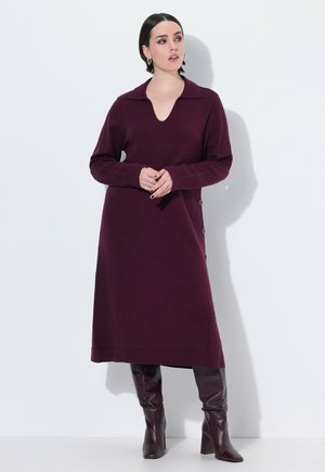 Abito lavorato a maglia bordeaux con colletto, scollatura a V e bottoni decorativi laterali. Indossato con stivali bordeaux lucidi fino al ginocchio. Texture liscia.