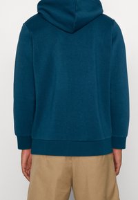 Sudadera con capucha en color azul verdoso, hecha de tela suave. Confeccionada con una cintura y puños acanalados, presenta un diseño en la parte posterior simple y sin costuras.
