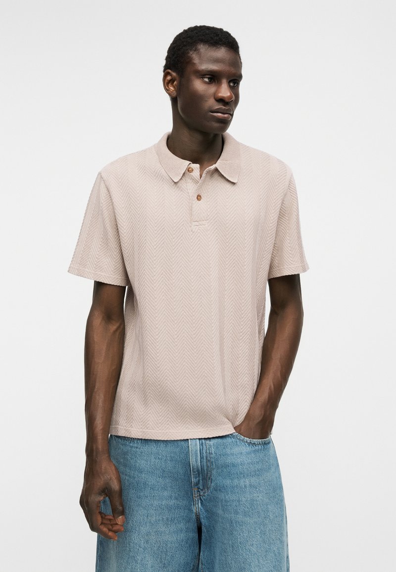 Homme noir portant un polo beige clair à manches courtes avec un motif en chevrons et un jean en denim bleu, debout avec une main dans la poche.
