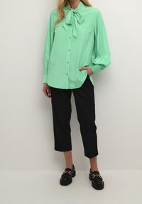 Blusa verde menta a maniche lunghe con colletto a fiocco, chiusura con bottoni e vestibilità rilassata; abbinata a pantaloni neri a tre quarti e mocassini.