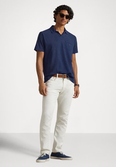 Polo de manga curta azul-marinho com gola e bolso, combinado com jeans brancos de corte slim e ténis azuis, posicionado contra um fundo simples.