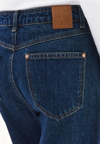 Jeans de mezclilla azul oscuro con una etiqueta de cuero que dice "OXXO," herrajes de cobre visibles y costuras naranjas en contraste en los bolsillos traseros.