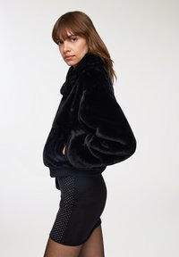 Giacca in pelliccia sintetica nera con colletto alto e tasche laterali. Polsi e orlo a coste. Indossata sopra una gonna nera con pois bianchi.