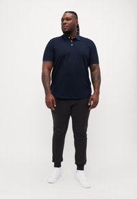 Mørkeblå polo skjorte med korte ermer, flad krave, og diskrete detaljer, i kombination med sorte tætsiddende joggere og hvide sneakers.