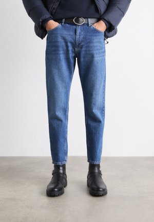 Jeans de mezclilla azul con corte ajustado y presillas para cinturón, combinados con botas negras hasta el tobillo. Una chaqueta oscura y acolchada es parcialmente visible en la parte superior.