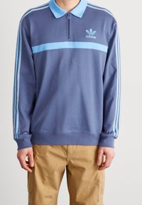 Modrý pulovr s límcem na zip, třemi světle modrými pruhy na rukávech a malým logem Adidas na hrudi. Vyrobený z měkké látky.