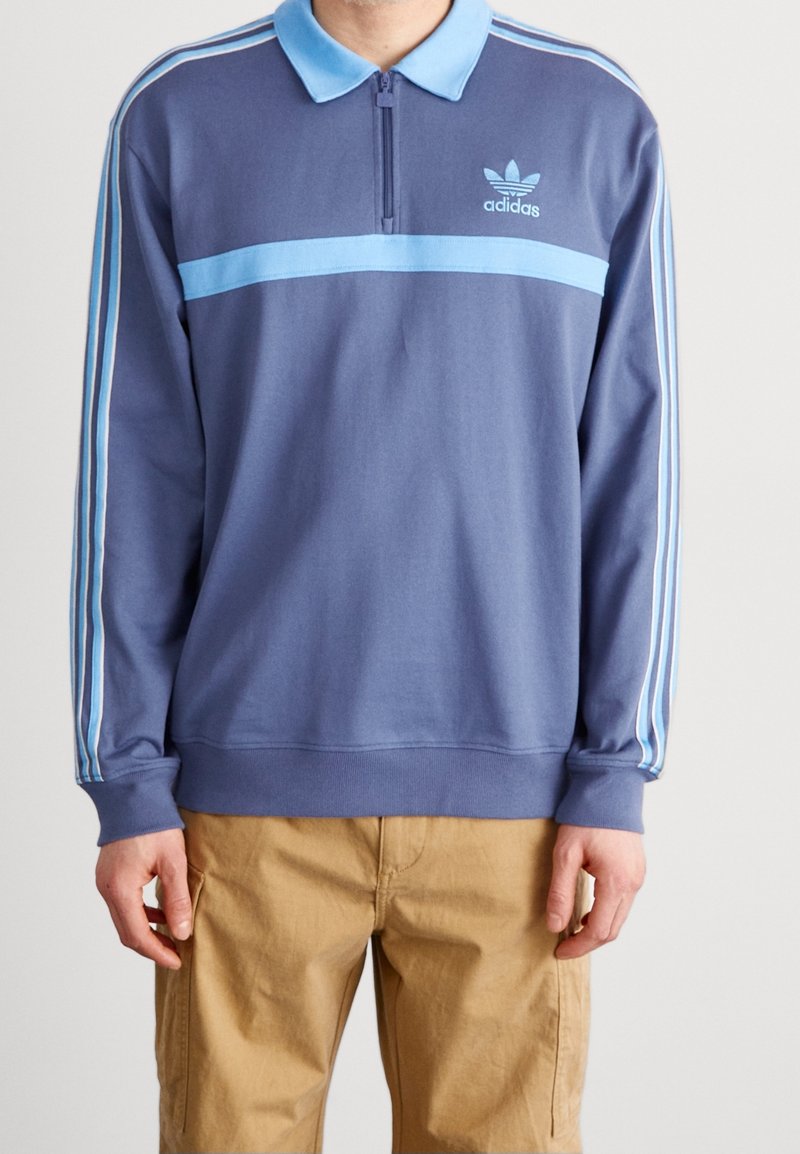 Modrý pulovr s límcem na zip, třemi světle modrými pruhy na rukávech a malým logem Adidas na hrudi. Vyrobený z měkké látky.
