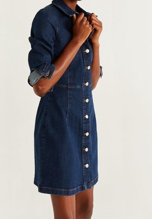 Femme portant une robe en jean bleu foncé avec boutons sur le devant et manches retroussées, tenant le col à deux mains devant un fond uni.