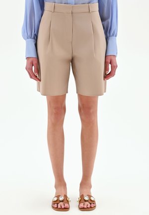 Pantaloncini beige su misura fino al ginocchio indossati con una camicia azzurro chiaro a maniche lunghe e sandali marroni adornati con grandi gemme rotonde.