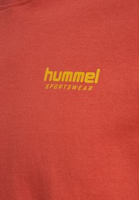 Raudonas medvilnės džemperis su geltonu "hummel" logo ir "SPORTSWEAR" užrašu. Lygus paviršius ir apvalia kaklo iškirpte.