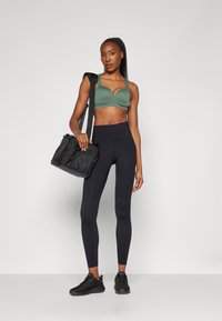 Grøn sportsbra, højtsiddende sorte leggings med perforerede detaljer, sorte sneakers og en sort duffeltaske med skulderrem.