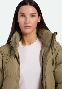 Chaqueta de plumón verde oliva con un alto cuello, cremallera frontal y cordones ajustables. Material texturizado y resistente al agua.
