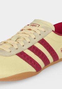 Κρεμ παπούτσι Adidas με κόκκινες υφασμάτινες ρίγες, μπεζ κορδόνια και λαστιχένια σόλα, εμφανίζεται σε κοντινή γωνία.