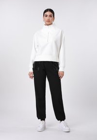 Λευκή φούτερ μπλούζα με ψηλό γιακά και φερμουάρ στο μπροστινό μέρος· συνδυασμένη με μαύρο παντελόνι φόρμας με παγιέτες στις τσέπες· λευκά αθλητικά παπούτσια.