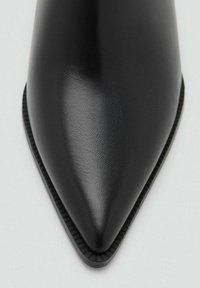 Zapato de tacón negro con punta afilada, hecho de cuero suave con superficie texturizada. Presenta una silueta esbelta y un borde cosido a lo largo de la suela.