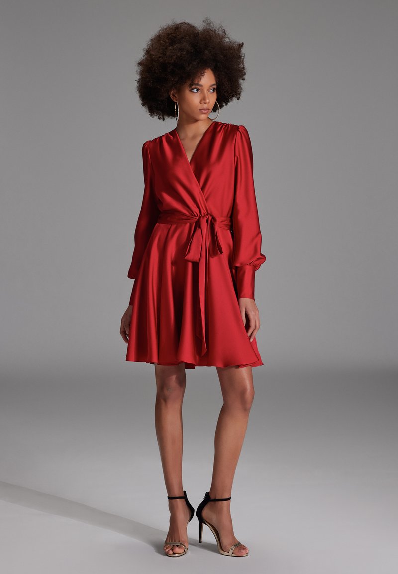 Robe portefeuille en satin rouge avec manches longues, ceinture à nouer, jupe évasée et décolleté en V. Associée à des sandales à talons hauts noires à lanières.